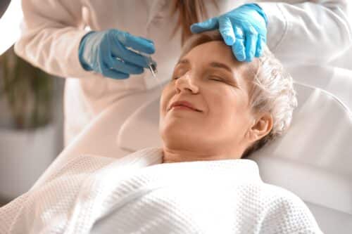 Mature woman receiving a facial filler or cosmetic injection while lying comfortably in a treatment chair, administered by a provider wearing blue medical gloves.